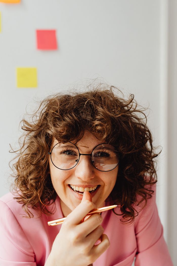 gallery-6 Curly-haired woman in glasses, wearing pink, smiling with pen in hand indoors.